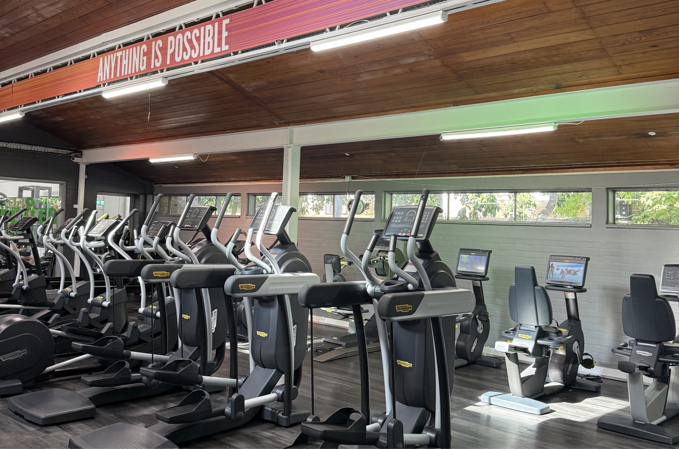 Gym floor at Lillie Road with rows of cardio machines under a wooden ceiling and a banner reading Anything is possible